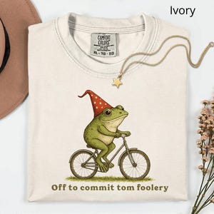 May include: Ivory-colored t-shirt featuring a whimsical illustration of a green frog wearing a red and white pointed hat, riding a bicycle. The text "Off to commit tom foolery" is printed below the frog. A gold star pendant necklace is draped across the shirt.