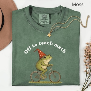May include: A moss green t-shirt featuring the phrase "Off to teach math" and a graphic of a frog on a bicycle wearing a wizard hat. A gold star pendant necklace is draped on the shirt.