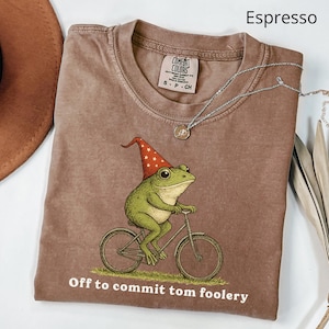 May include: Espresso-colored t-shirt featuring a green frog riding a bicycle and wearing a red and white pointed hat. The text "Off to commit tom foolery" is printed below the frog. A silver necklace with a pendant is draped across the shirt.