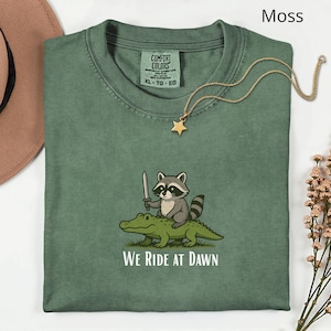 Pu&ograve; includere: Una maglietta verde muschio con una grafica di un procione con una spada che cavalca un alligatore, con la scritta "WE RIDE AT DAWN". Una collana con un ciondolo a stella dorata &egrave; drappeggiata sulla maglietta.