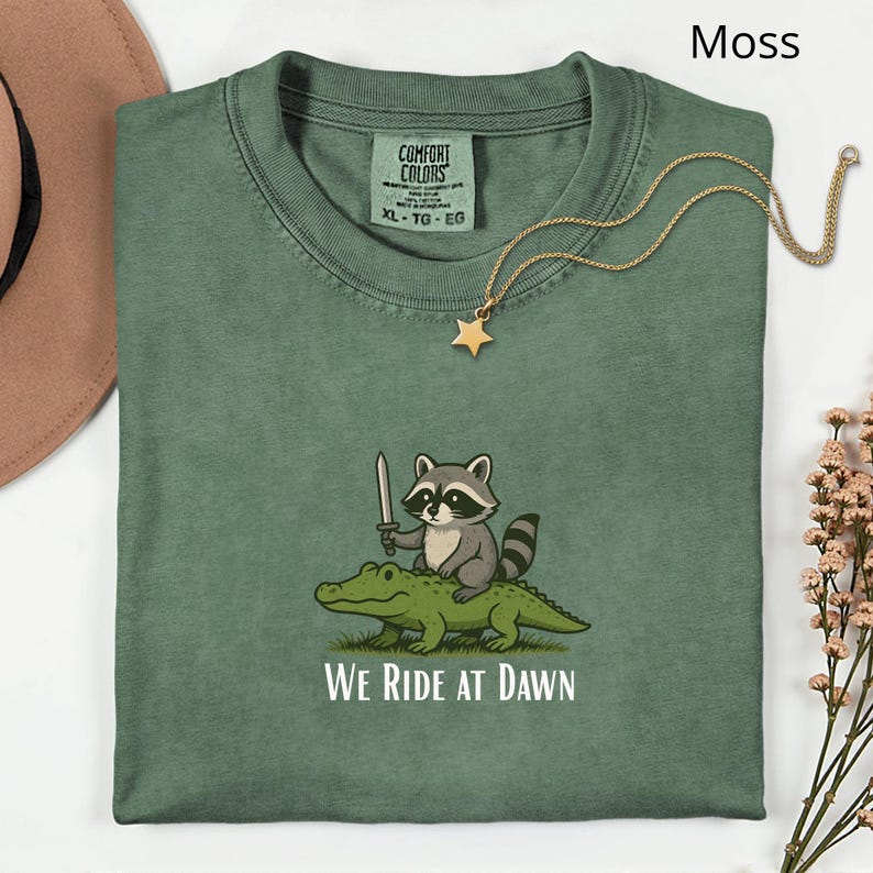 Pu&ograve; includere: Una maglietta verde muschio con una grafica di un procione con una spada che cavalca un alligatore, con la scritta "WE RIDE AT DAWN". Una collana con un ciondolo a stella dorata &egrave; drappeggiata sulla maglietta.