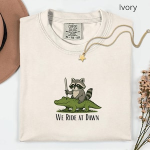 Pu&ograve; includere: T-shirt color avorio con grafica di un procione con una spada che cavalca un alligatore. Il testo "WE RIDE AT DAWN" &egrave; sotto la grafica. Una collana dorata con un ciondolo a stella &egrave; drappeggiata sulla maglietta.