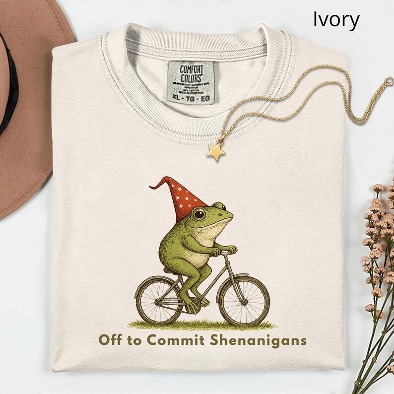 May include: Ivory-colored t-shirt featuring a green frog wearing a red hat with white stars, riding a bicycle. The text "Off to Commit Shenanigans" is printed below the frog. A gold necklace with a star pendant is draped on the shirt.