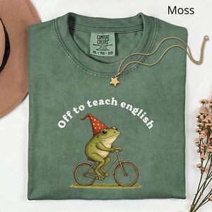 May include: A moss green t-shirt featuring the phrase "Off to teach english" and a frog riding a bicycle, wearing a red and white hat. A gold star pendant necklace is draped across the shirt.