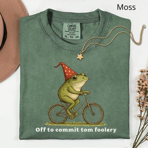 May include: Moss green t-shirt featuring a frog on a bicycle, wearing a red and white hat. The text "Off to commit tom foolery" is printed below the graphic. A gold star pendant necklace is draped on the shirt.