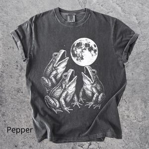 May include: Pepper-colored t-shirt featuring a white graphic of three frogs singing under a full moon. The frogs are detailed with open mouths and textured skin. The shirt has rolled-up sleeves and a crew neck.