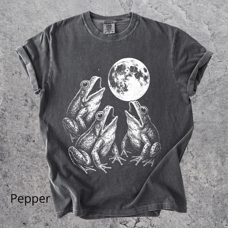 May include: Pepper-colored t-shirt featuring a white graphic of three frogs singing under a full moon. The frogs are detailed with open mouths and textured skin. The shirt has rolled-up sleeves and a crew neck.