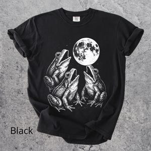 May include: Black t-shirt featuring a detailed white illustration of three frogs singing under a full moon. The shirt has a classic crew neck and short sleeves. The word "Black" is printed at the bottom of the shirt.