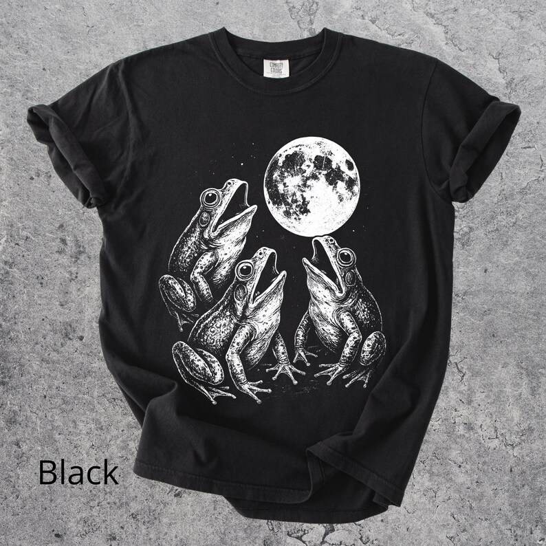 May include: Black t-shirt featuring a detailed white illustration of three frogs singing under a full moon. The shirt has a classic crew neck and short sleeves. The word "Black" is printed at the bottom of the shirt.