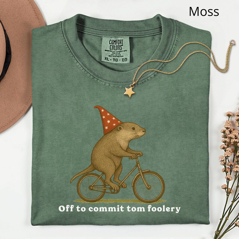 May include: Moss green t-shirt with a graphic of a rodent wearing a wizard hat and riding a bicycle. The text "Off to commit tom foolery" is below the graphic. A gold star pendant necklace is on the shirt.