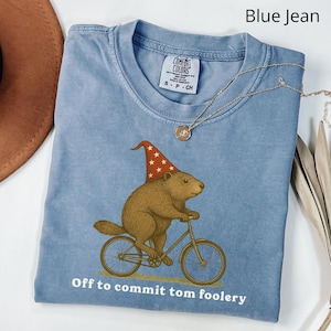 Op de afbeelding: Blauw spijkerbroekkleurig T-shirt met een afbeelding van een bruine bever met een rood-witte sterrenhoed, fietsend op een fiets. De tekst "Off to commit tom foolery" staat eronder. Een gouden ketting ligt over het shirt.