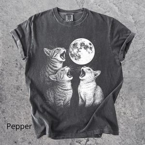 May include: Dark grey t-shirt with a graphic of three cats howling at a full moon. The cats are in a detailed, black and white illustration. The word "Pepper" is printed at the bottom left of the shirt.