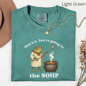 May include: A light green t-shirt with the text "That's it. You're going in the SOUP" and a cartoon frog chef stirring a pot of soup. A gold star pendant necklace is draped on the shirt.