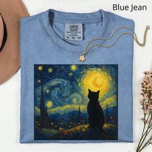 May include: A blue jean coloured t-shirt featuring a graphic of a black cat against a swirling night sky, inspired by Van Gogh's "Starry Night." A gold star pendant necklace is on the shirt. The shirt has the text "Comfort Colors" and size XL-TG-EG.