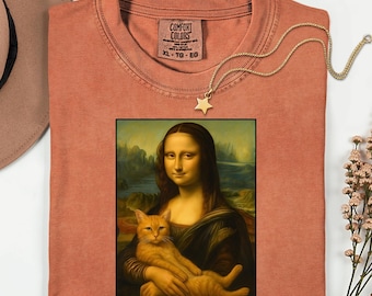 Peut inclure: Un t-shirt couleur corail avec une image encadrée de la Joconde tenant un chat. Un collier pendentif étoile en or est posé sur le t-shirt. Le t-shirt porte une étiquette Comfort Colors et est étiqueté XL-TG-EG.