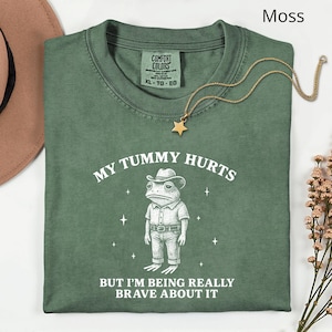 Op de afbeelding: Een mosgroen t-shirt met de tekst "MY TUMMY HURTS BUT I'M BEING REALLY BRAVE ABOUT IT" en een cartoon kikker met een cowboyhoed. Een gouden sterrenketting ligt over het shirt.