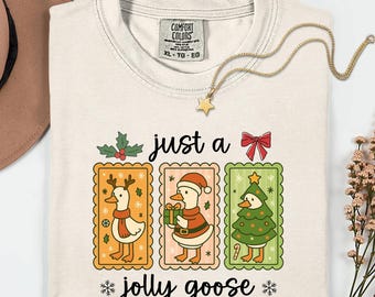 May include: Ivory-colored t-shirt with a festive "just a jolly goose" design. The graphic features three cartoon geese dressed in holiday attire, each within a decorative frame. A gold necklace with a star pendant rests on the shirt.
