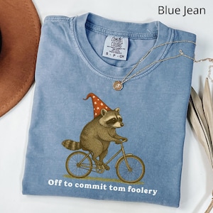 Könnte beinhalten: Hellblaues T-Shirt mit einem Waschbären auf einem Fahrrad, der einen rot-weißen Hut trägt. Der Text "Off to commit tom foolery" ist unten aufgedruckt. Eine goldene Halskette mit Anhänger ist ebenfalls sichtbar.