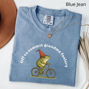 May include: A blue jean colored t-shirt with the phrase "Off to commit grandma foolery" encircling a graphic of a frog riding a bicycle. The frog wears a red and white hat. A gold necklace is draped across the shirt.