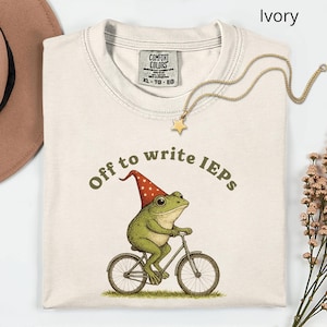Könnte beinhalten: Elfenbeinfarbenes T-Shirt mit dem Text "Off to write IEPs" und einer Grafik einer Frosch auf einem Fahrrad, der einen rot-weißen Hut trägt. Eine goldene Sternkette liegt über dem Shirt. Das Shirt ist ein Comfort Colors XL-TG-EG.