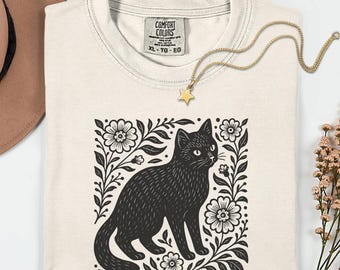 Può includere: T-shirt color avorio con un'illustrazione di un gatto nero circondato da elementi floreali. La maglietta ha un girocollo e maniche corte. Una collana color oro con un ciondolo a forma di stella è drappeggiata sulla maglietta. La maglietta è etichettata XL-TG-EG.