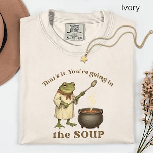 May include: Ivory-colored t-shirt with a frog chef graphic holding a spoon over a pot of soup. The text reads "That's it. You're going in the SOUP." A gold star necklace is draped on the shirt.