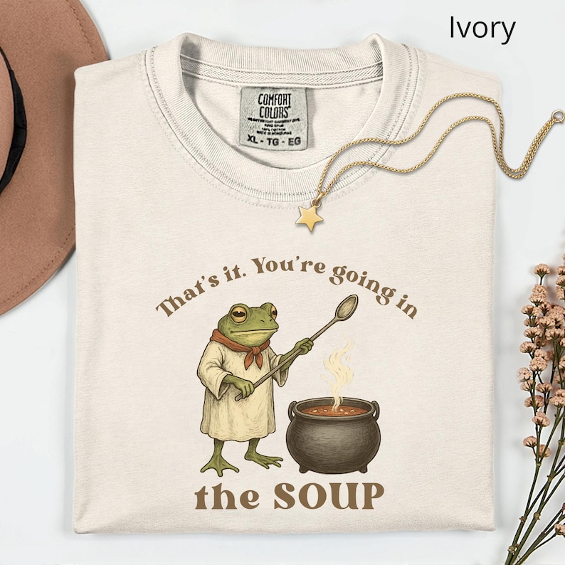 May include: Ivory-colored t-shirt with a frog chef graphic holding a spoon over a pot of soup. The text reads "That's it. You're going in the SOUP." A gold star necklace is draped on the shirt.