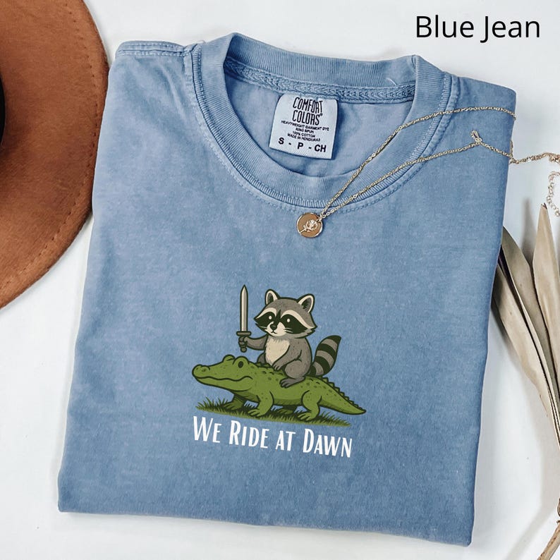Pu&ograve; includere: Una maglietta blu jeans con un procione dei cartoni animati che cavalca un alligatore e tiene una spada. Il testo "WE RIDE AT DAWN" &egrave; stampato sotto la grafica. Una collana d'oro con un ciondolo &egrave; drappeggiata sulla maglietta.