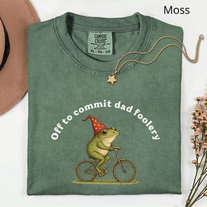 May include: A moss green t-shirt with the text "Off to commit dad foolery" surrounding a cartoon frog riding a bicycle. The frog wears a red and white pointed hat. A gold star pendant necklace is draped across the shirt.