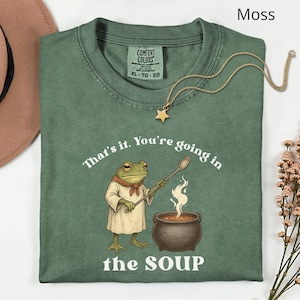 May include: Moss green t-shirt with the text "That's it. You're going in the SOUP." A cartoon frog wearing a chef's outfit stirs a pot of soup. A gold star pendant necklace is draped on the shirt.