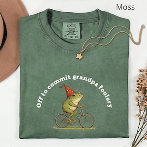 May include: Moss green t-shirt with a frog riding a bicycle, wearing a red and white hat. The text "Off to commit grandpa foolery" is arched above the frog. A gold star pendant necklace is on the shirt.