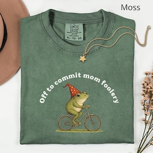 Puede incluir: Camiseta verde musgo con el texto "Off to commit mom foolery" y una rana de dibujos animados en bicicleta, con un sombrero rojo y blanco. También se ven un collar con colgante de estrella dorada y un sombrero marrón.