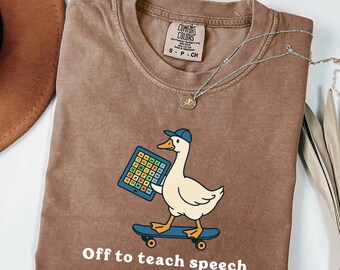 May include: Espresso-colored t-shirt with a graphic of a duck wearing a cap and holding a tablet while skateboarding. The text "Off to teach speech" is printed below the graphic. A silver necklace is draped across the shirt.