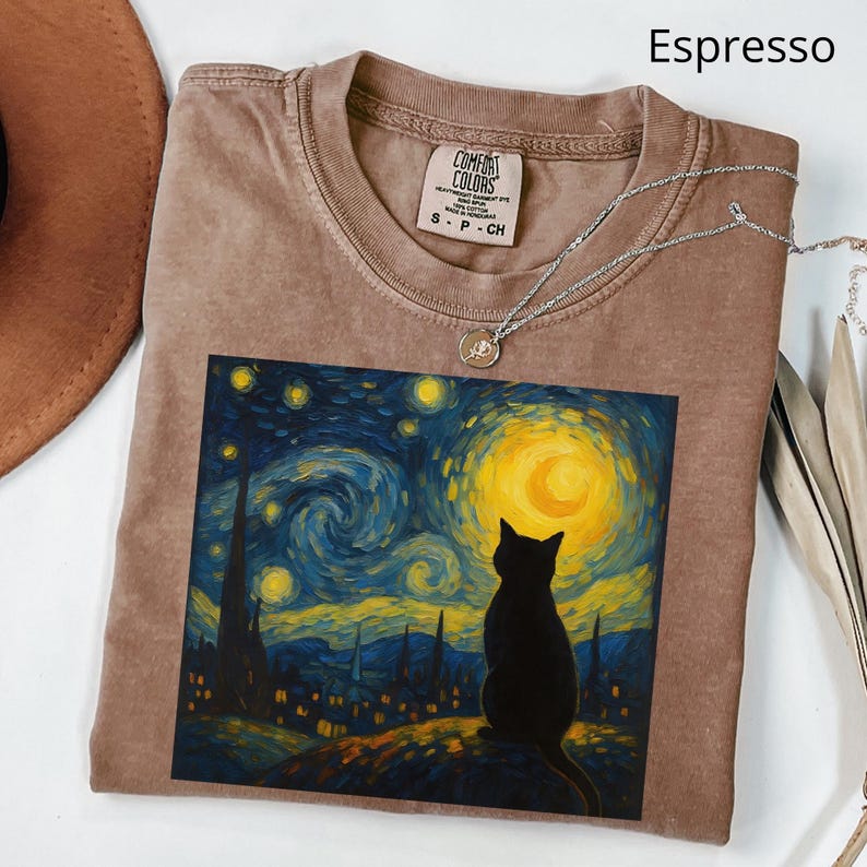 May include: Espresso-colored t-shirt featuring a graphic of a black cat silhouetted against a starry night sky, reminiscent of Van Gogh's style. The shirt has a crew neck and short sleeves. A silver necklace with a small pendant is draped across the shirt.
