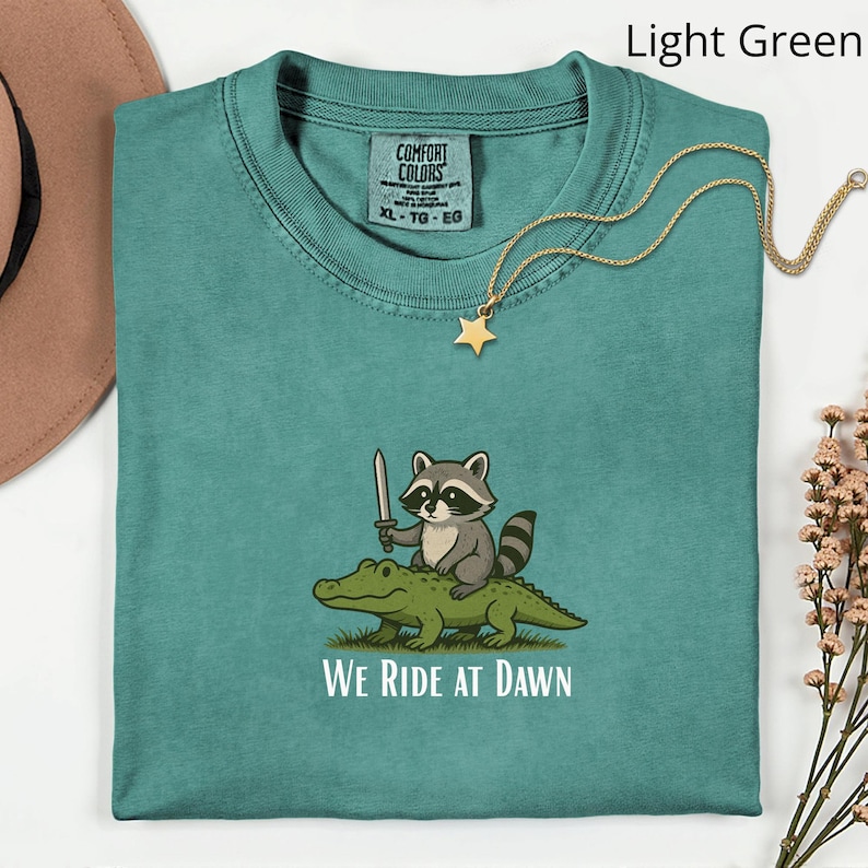 Pu&ograve; includere: T-shirt verde chiaro XL Comfort Colors con una grafica di un procione con una spada che cavalca un alligatore, con la scritta "WE RIDE AT DAWN". Una collana con pendente a stella dorata &egrave; drappeggiata sulla maglietta.