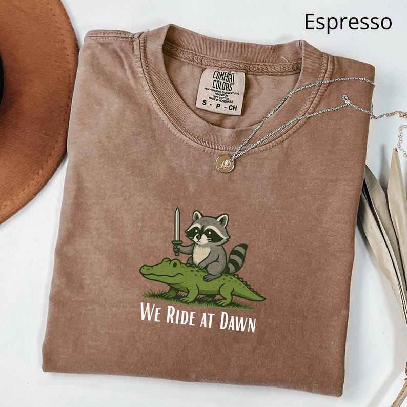 Pu&ograve; includere: T-shirt color espresso con una grafica di un procione con una spada che cavalca un alligatore. Il testo "WE RIDE AT DAWN" &egrave; sotto la grafica. Una collana d'argento con un ciondolo &egrave; sulla maglietta.