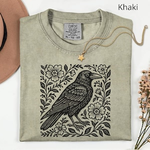 May include: Khaki-colored t-shirt with a black raven illustration surrounded by floral elements. The shirt has a crew neck and short sleeves. A gold necklace with a star pendant rests on the shirt. The shirt is labeled XL-TG-EG.