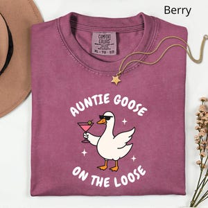 May include: Berry-colored t-shirt with the text "Auntie Goose on the Loose" and a cartoon goose wearing sunglasses and holding a cocktail. A gold necklace with a star pendant is draped across the shirt.