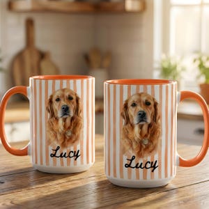 Op de afbeelding: Twee keramische mokken met een verticaal oranje en wit gestreept patroon. Elke mok toont een portret van een golden retriever en de naam Lucy in zwart cursief schrift. De mokken hebben een oranje handvat en een oranje binnenkant.