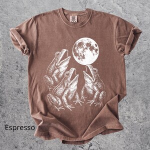 May include: Espresso-colored t-shirt featuring a white illustration of three frogs singing under a full moon. The shirt has a classic crew neck and short sleeves. The word "Espresso" is printed at the bottom left.