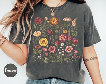 May include: Dark grey t-shirt featuring a colorful floral design with various flowers in shades of red, yellow, pink, and white. The shirt has a crew neck and short sleeves. The word "Pepper" is printed on a circle.