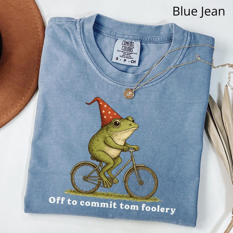 May include: A light blue t-shirt with a whimsical illustration of a green frog wearing a red and white pointed hat, riding a bicycle. The text "Off to commit tom foolery" is printed below the image. A silver necklace is draped across the shirt.