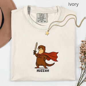 May include: Ivory-colored t-shirt featuring a cartoon otter with a sword and cape, the word "Huzzah" below. The shirt has a Comfort Colors tag and a gold star necklace. The shirt is folded on a white surface.