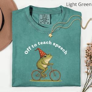 Op de afbeelding: Lichtgroen t-shirt met de tekst "Off to teach speech" en een kikker op een fiets. De kikker draagt een rood-witte hoed. Een gouden sterrenketting ligt op het shirt.