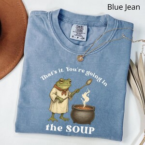May include: Blue jean colored t-shirt with the text "That's it. You're going in the SOUP." A cartoon frog wearing a chef's outfit stirs a pot of soup. A gold necklace is draped across the shirt.