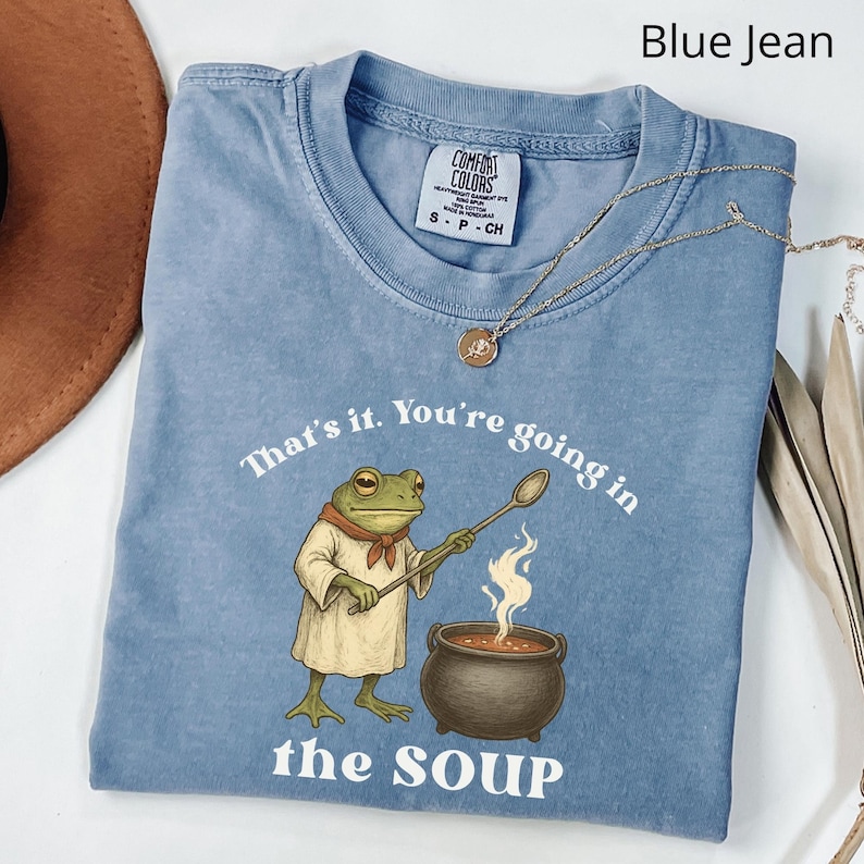 May include: Blue jean colored t-shirt with the text "That's it. You're going in the SOUP." A cartoon frog wearing a chef's outfit stirs a pot of soup. A gold necklace is draped across the shirt.