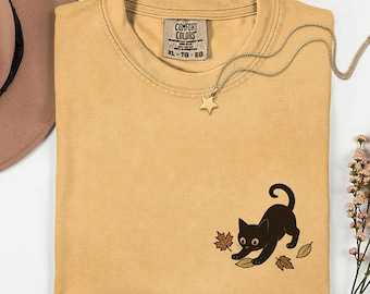 Puede incluir: Camiseta color mostaza con un gráfico de un gato negro jugando con hojas de otoño. La camiseta tiene una etiqueta Comfort Colors y un collar con colgante de estrella dorada. La camiseta está doblada.