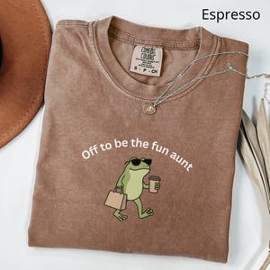 May include: Espresso-colored t-shirt with the phrase "Off to be the fun aunt" and a cartoon frog wearing sunglasses and carrying a shopping bag and coffee cup. A silver necklace with a pendant is draped across the shirt.