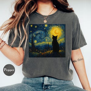 May include: Dark grey t-shirt featuring a graphic of a black cat silhouette against a starry night sky with a yellow moon. The artwork is reminiscent of Van Gogh's style. The shirt has a crew neck and short sleeves.