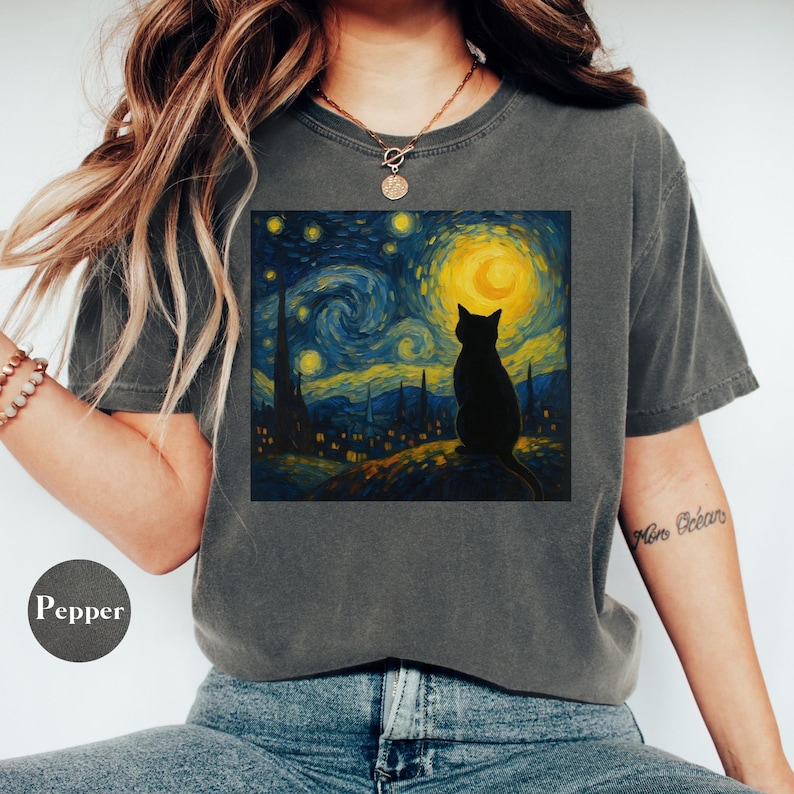 May include: Dark grey t-shirt featuring a graphic of a black cat silhouette against a starry night sky with a yellow moon. The artwork is reminiscent of Van Gogh's style. The shirt has a crew neck and short sleeves.